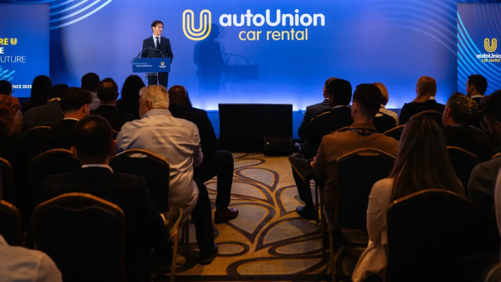 AutoUnion COO speaks to audience in front of wide blue screen with company logo on stage.