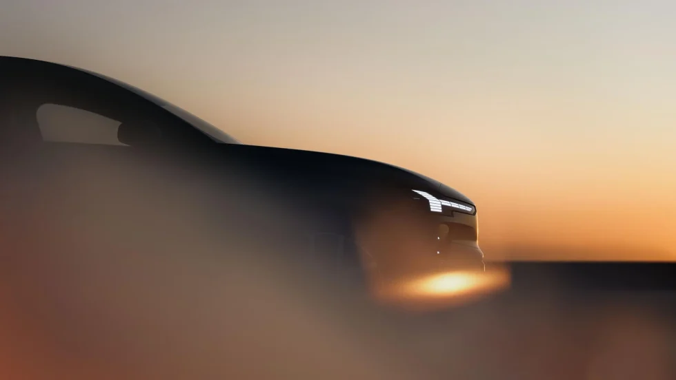 A Volvo EX60 drives on a gravel road with woody surrounding it