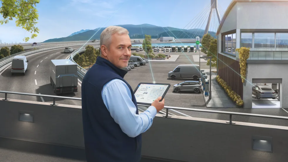 Man using tablet to monitor connected fleet vehicles and EV chargers near highway, representing Bosch’s AI-driven predictive maintenance and Uptake acquisition.