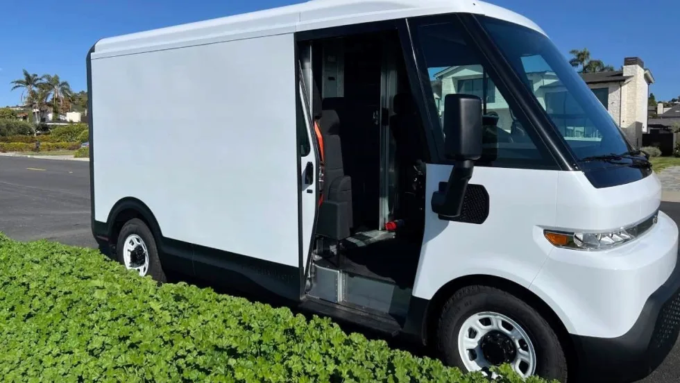 A white electric delivery van parked along a residential curb with low green shrubs.
