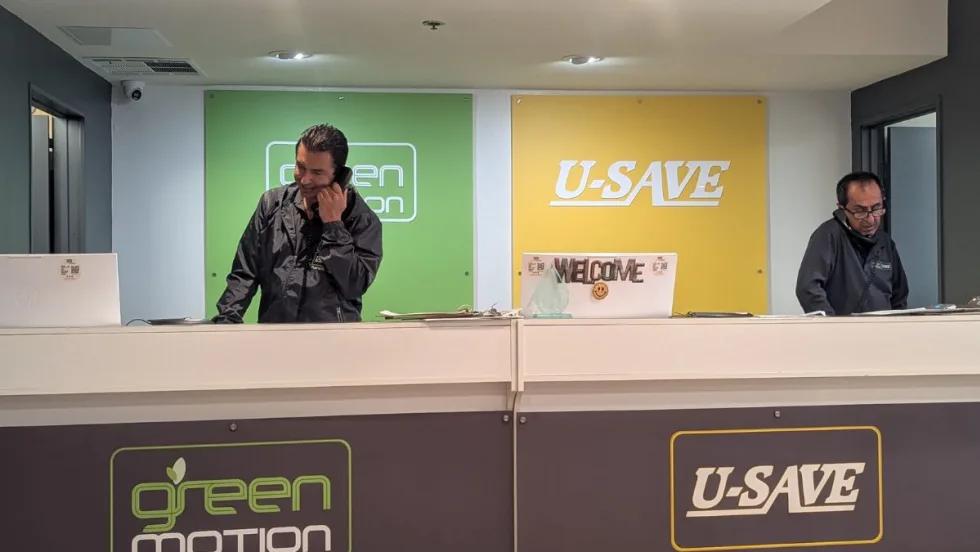 Two men working at a Green Motion U-Save car rental county with green and yellow logo backdrops.