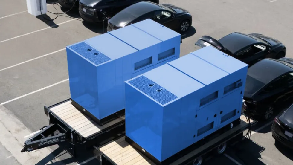 Two blue large box mobile chargers parked along a row of black Tesla EV sedans in a parking lot.
