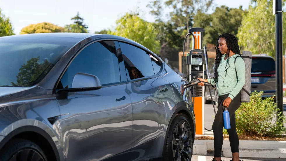 Person charging an electric vehicle at a public EV charging station, holding the charging connector next to a parked electric car outdoors.