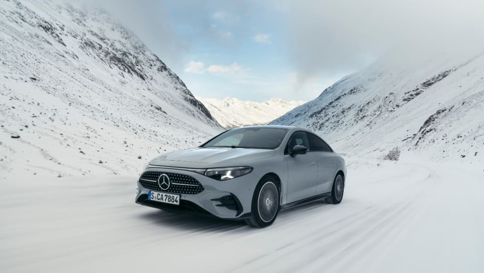 A metallic white hybrid Mercedes-Benz CLA drives between two snowy mountains.