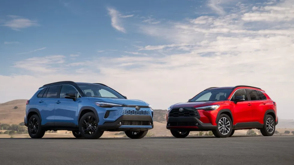 The 2026 Toyota Corolla Cross and its hybrid counterpart sit next to each other on the road with a blue sky background behind them. 