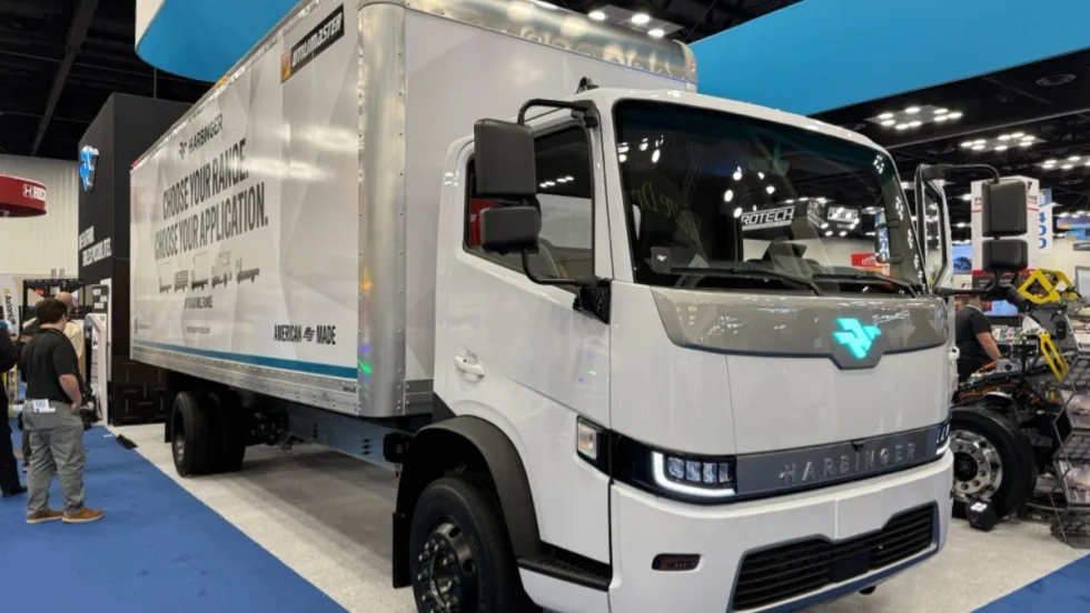 Harbinger HC Series Cab medium-duty electric work truck driving through a city with glass office towers, demonstrating the low cab forward fleet vehicle platform introduced at Work Truck Week.