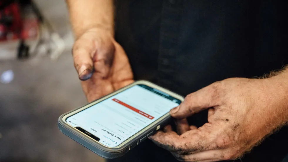  Grease-stained hands hold a smartphone displaying a digital fleet maintenance or vehicle service form.