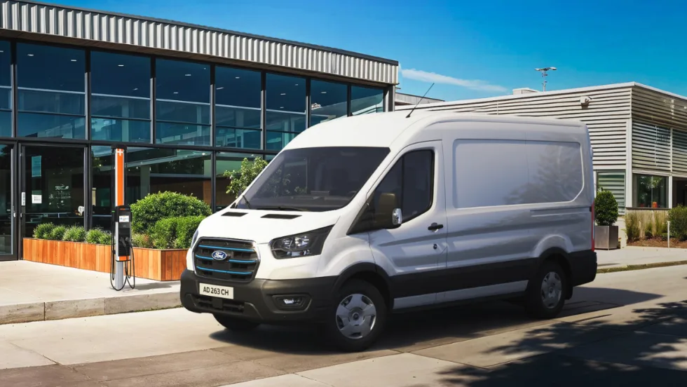 White Ford electric cargo van parked and charging at a ChargePoint station outside a modern commercial building.