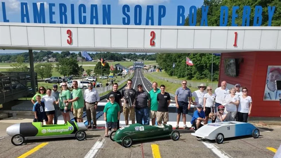 A group of participants and organizers stand behind three colorful soap box cars at the starting line of the All-American Soap Box Derby track in Akron, Ohio, celebrating the launch.