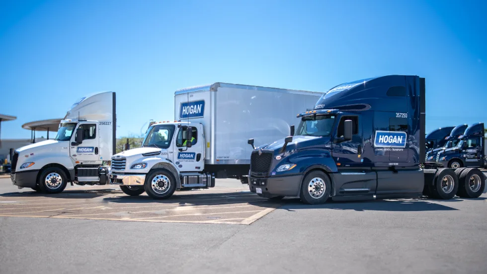 Three Hogan semi-trucks sit in a row in a parking lot on a sunny day.