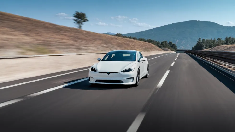 A white Tesla Model S driving down a paved road with hills and mountain in background on a sunny day.