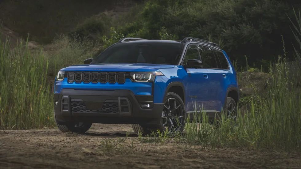 A blue Jeep Cherokee SUV parked outdoors on a dirt trail is surrounded by tall grass and greenery.