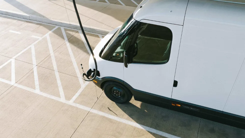 A white Rivian commercial van is charging in a parking lot.