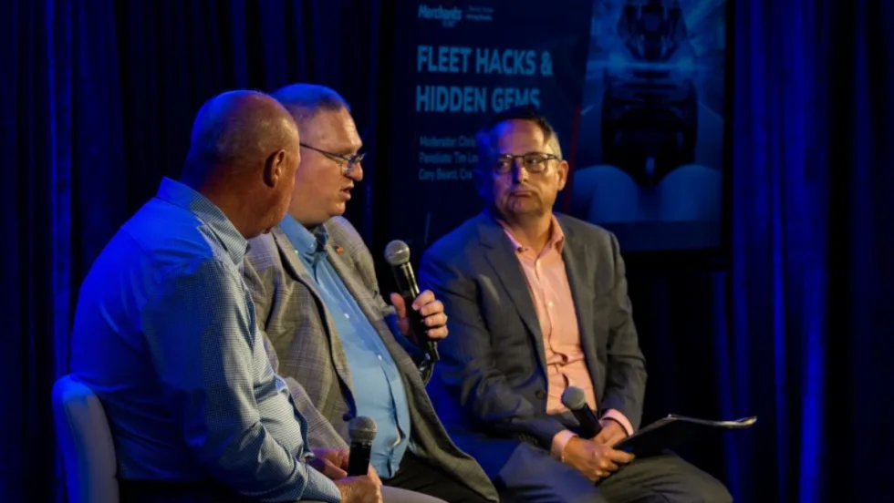 Cory Beard holds a mic next to Tim Lovett and Chris Brown, who sit on a stage for a panel discussion at the Merchant's Fleet conference in New Hampshire.