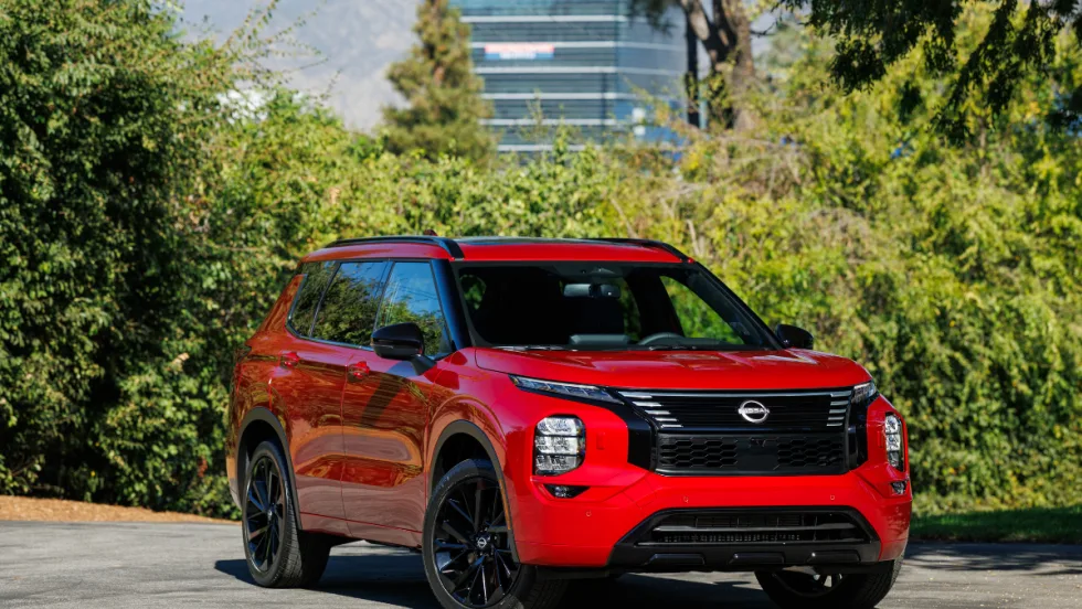 A red 2026 Nissan Rogue plug-in hybrid drives on a road next to bushes.