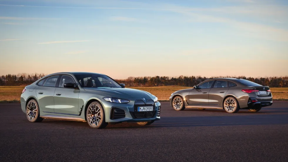 Two 2025 BMW i4 series cars (one light blue and one dark gray) parking on pavement with a field and trees in the background.