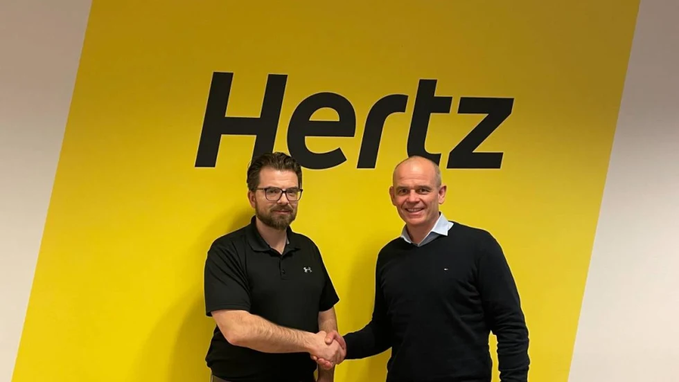 Two executives shaking hands in front of bold yellow Hertz sign.