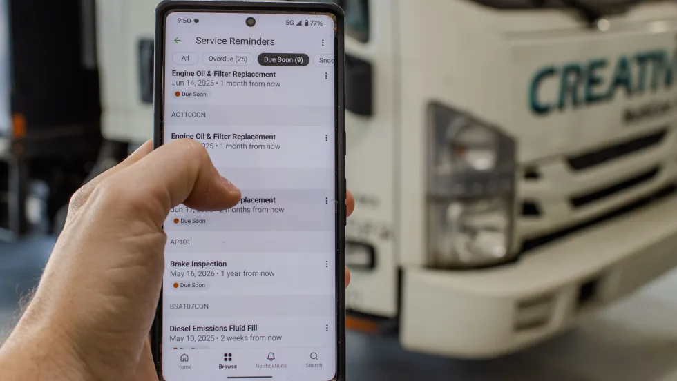 A person holds a smartphone displaying a list of vehicle service reminders, including engine oil and filter replacement, while standing in front of a white commercial truck.