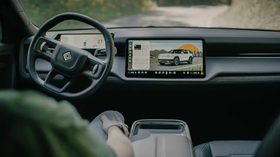 Interior view of a Rivian vehicle showing steering wheel and digital display screen with vehicle interface, representing autonomous technology development.
