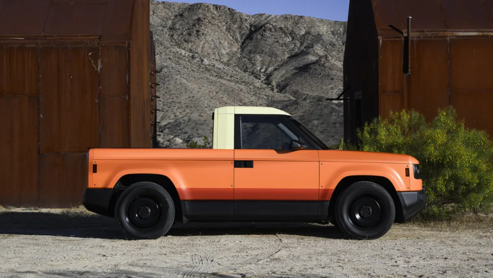 Orange Slate electric pickup truck parked outdoors between industrial structures, representing the company’s upcoming customizable EV supported by new funding.