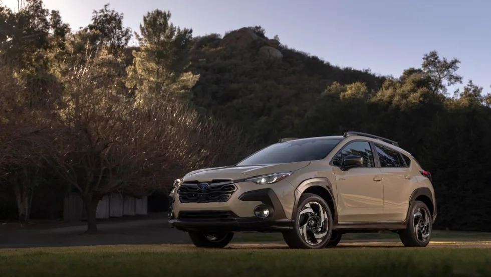 “A beige Subaru Crosstrek SUV parked on grass, surrounded by trees and hills under a clear sky at sunset.