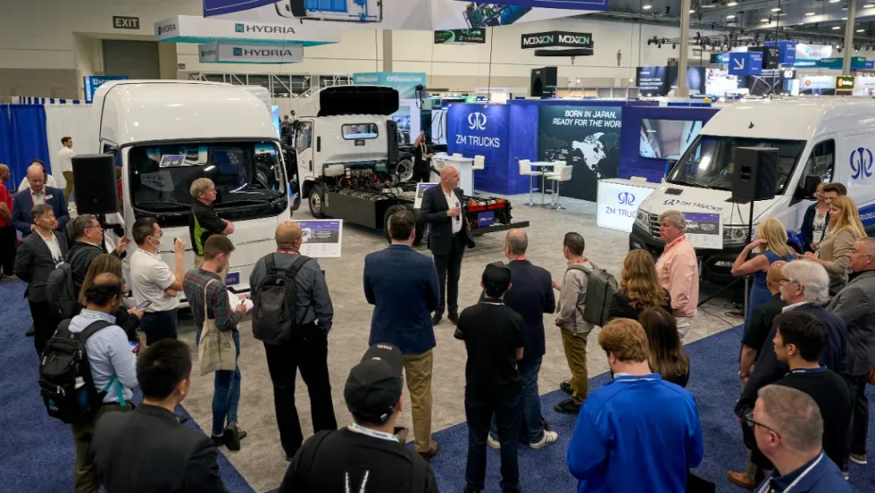 Media gather on show floor to listen to a press conference, trucks in background. 