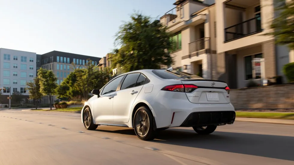 A 2026 Toyota Corolla drives on the road towards a glass building.