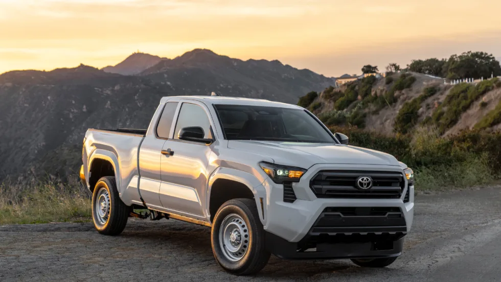 A white 2026 Toyota Tacoma sits in a sunset landscape with mountains behind the road.