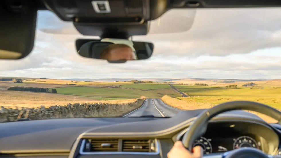 A car renter driving a right-side steering wheel on the left side of a two-lane road.