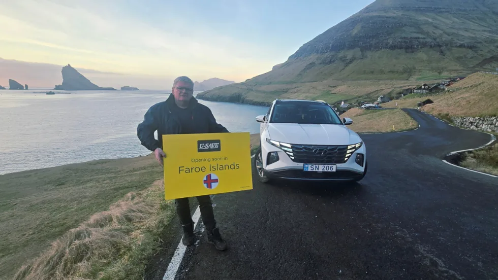 U-Save franchisee holding a promotional sign in front of a white rental car along the rugged coastline of the Faroe Islands. 