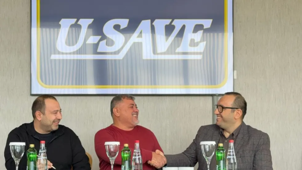 Three U-Save franchise operators are congratulating each other along a horizontal table beneath a large framed U-Save logo print.
