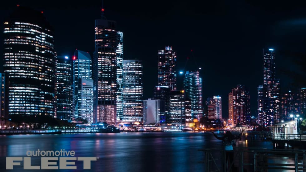 Nighttime view of a modern city skyline reflected in the water, with brightly lit skyscrapers and a silhouetted figure standing on a dock in the foreground.