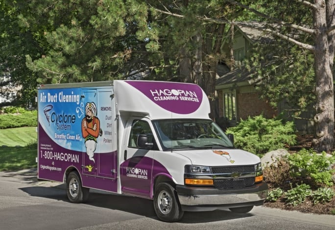 Purple-and-white Hagopian Cleaning Services box truck drives along a tree-lined residential street.