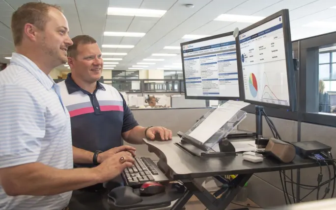 Two fleet employees review vehicle data, charts, and maintenance information displayed on dual computer monitors in an office.