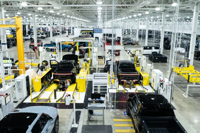 Rivian vehicle assembly line inside a large manufacturing plant with multiple electric trucks being built.