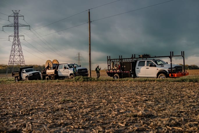 A lineup of heavy-duty trucks equipped with utility upfits parked in an open field beneath power lines, ready for deployment to support disaster recovery efforts.