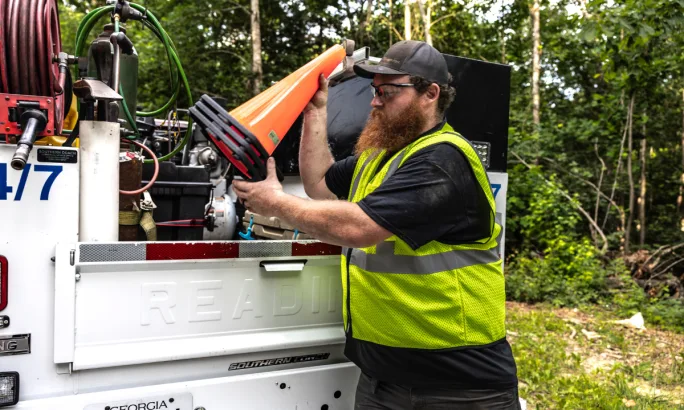 Man in high-visibility safety vest unloading a stack of orange traffic cones from a work truck equipped with tools and equipment, surrounded by trees.