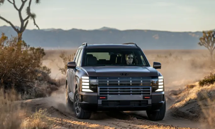 Front view of the 2026 Hyundai Palisade XRT PRO driving on a rugged desert trail, kicking up dust with the driver visible inside, set against a mountainous background.