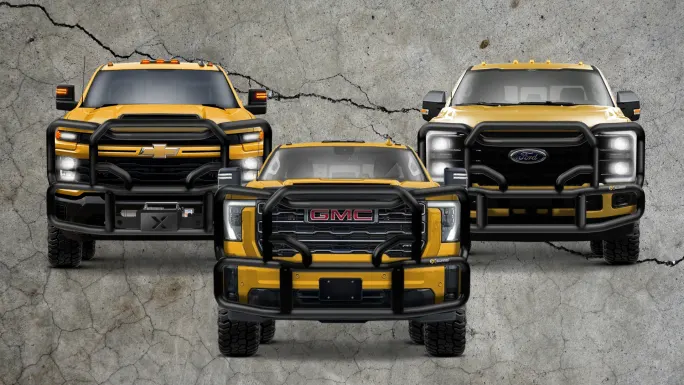 Three yellow heavy-duty trucks (Chevrolet, GMC, and Ford) with Ex-Guard grille guards, parked against a cracked concrete background.