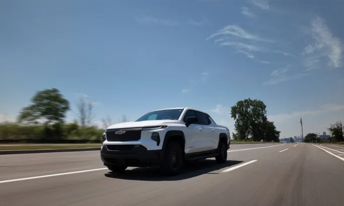 A white Chevrolet Silverado EV Max Range Work Truck drives on a sunny day during a range test, with trees, an American flag, and a distant city skyline in the background.