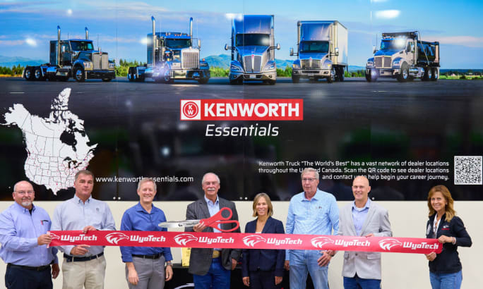 Group of business people stand in front of a ribbon about to be cut with signage behind them for Kenworth showing multiple trucks.