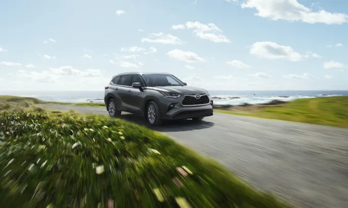 Gray Toyota Highlander SUV driving on coastal road with green grass and ocean background.