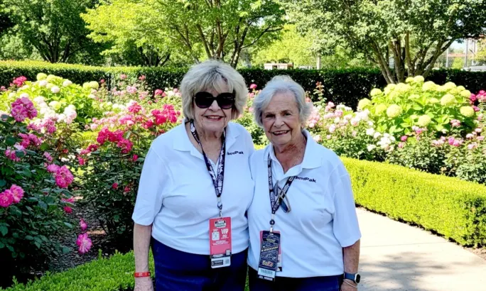 Two women, Ginger Henthorn and Carolyn Strelau, smiling in a flower garden while wearing white BendPak shirts and event passes.