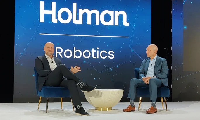 Two businessmen discuss robotics on stage during a conference wit.h Holman Robotics logo in the background