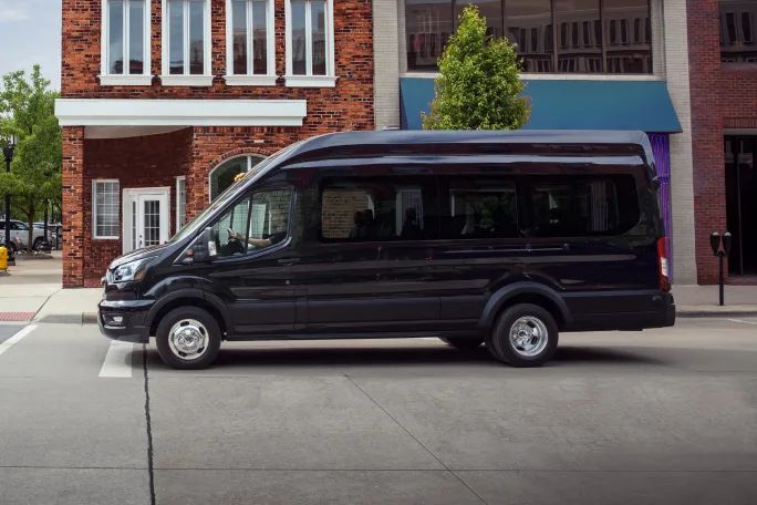 Side view of a 2026 Ford Transit Passenger Van driving in an urban area. Image highlights the updated model’s safety tech, software upgrades and configuration options for passenger and shuttle fleets.