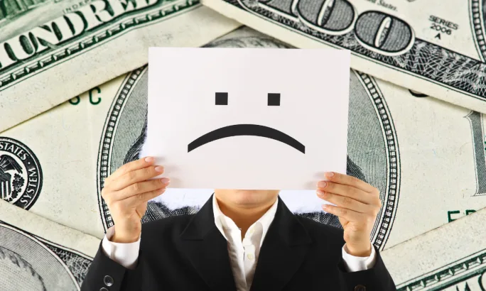 Person in a suit holding a paper with a sad face over their own face, standing in front of a backdrop of hundred-dollar bills, symbolizing financial strain.