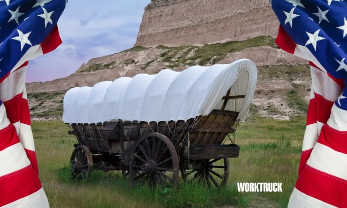 Covered wagon framed by American flags in a grassy landscape, representing early freight transport and the origins of American work vehicles.