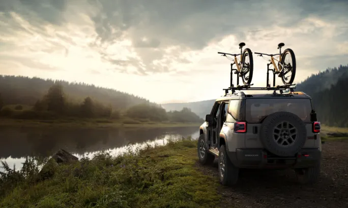 Rear view of a Jeep Recon parked by a lake at sunrise with two bikes on the roof rack.