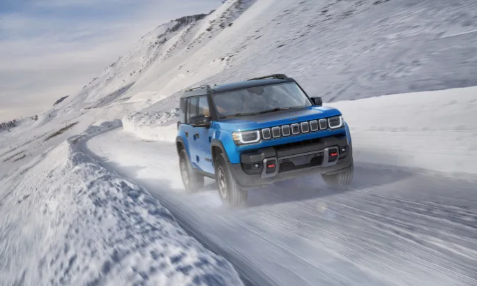 Blue 2026 Jeep Recon driving on a snowy mountain road, kicking up snow.