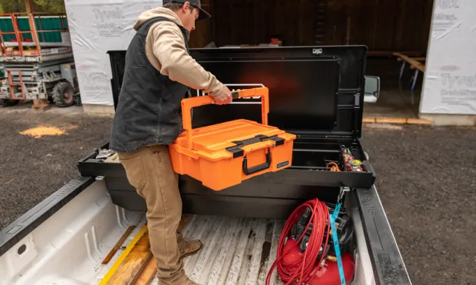 Worker lifts an orange DECKED case into a rear-mounted DECKED truck-bed tool box with the lid open.